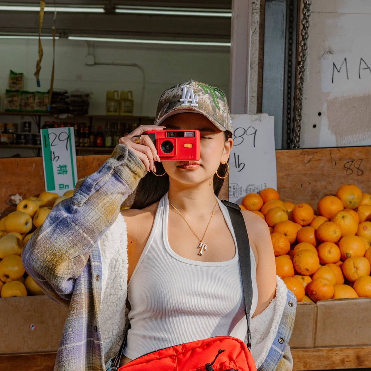 A woman in a street market setting wearing a red retro-style camera, a camouflage LA cap, a white ribbed tank top, a blue and white plaid shirt, and a red crossbody bag, with oranges and pears in the background.

Long Weekend 35mm Reusable Film Camera Analog