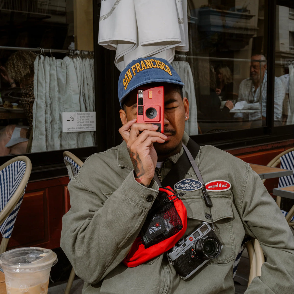 A man sitting at an outdoor cafe wearing a blue 'San Francisco' cap, a green utility jacket, and a red crossbody bag holding a red instant camera. A vintage camera hangs from his chest pocket.

Long Weekend 35mm Reusable Film Camera Analog