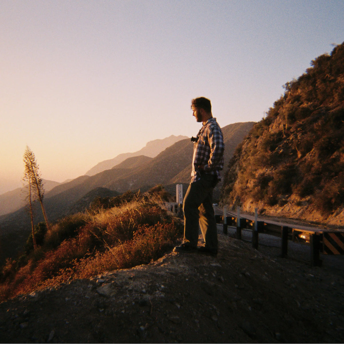 A man in a plaid shirt and dark pants stands on a dirt road overlooking a mountain landscape at sunset, with dry brush, a guardrail, and layered hills in the background.

Long Weekend 35mm Reusable Film Camera Analog